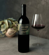 A bottle of Paul Hobbs Coombsville Napa Valley Cabernet Sauvignon 2019 stands on a gray surface beside a glass of red wine, with a potted plant in the background.