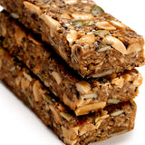 Three stacked Peanut Butter Ivan Bars filled with roasted peanuts, superfood seeds, and grains display a chewy texture and glossy surface against a white background.