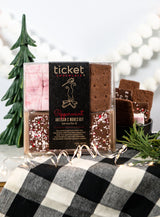 A Peppermint Artisan S'mores Kit featuring gourmet marshmallows and artisan chocolate bars is displayed on a black and white plaid cloth, surrounded by holiday decorations, a small tree, and festive string lights.