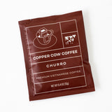 A brown packet labeled Single-Serve Pour Over Churro Coffee features a coffee cup, milk carton, cow graphic, and "Premium Vietnamese Coffee" text. Enjoy this convenient treat. Net weight: 0.4 oz (12g).