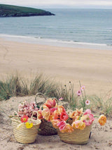 Three Petite Market Baskets, brimming with colorful flowers, sit on sandy grass overlooking a calm beach and ocean.