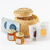 The Petite Tea Gift Basket is shown partially open on a white background, revealing straw filling. Displayed beside it are a Honey Sampler, two Olive Leaf Tea Bags, and two shortbread cookie packets.