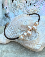 The Pink Pearl Cluster Reflection Bracelet features five pink pearls on leather, shown on a seashell with an iridescent backdrop.