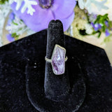 The Raw Amethyst Statement Ring, featuring a hand-crafted geometric purple gemstone in sterling silver, rests on a black velvet stand. Soft, blurred purple and white flowers enhance the dreamy ambiance.