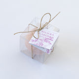 The Riviera Gardens Bath Bomb, pink with rose kaolin clay, is packaged in a clear plastic box tied with brown twine and labeled with a purple floral design. It sits on a white background.