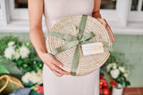 A person in a light dress holds a Round African Basket with a Lid tied with a green ribbon and a Santa Barbara Company tag. Flowers and greenery blur in the background, showcasing this handcrafted decorative piece.