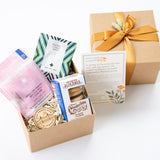 A gift box with an assortment of artisan snacks and treats, including packaged gluten-free alfajores cookies, rose petal caramels, and Pacific mint tea with teabags, accompanied by a San Francisco wood magnet, all displayed with a bow and information cards.