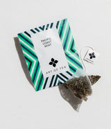 A packet labeled “Pacific Coast Mint” by Art of Tea rests on a white surface, with a pyramid tea bag and an “ENJOY THE CUP” tag—a perfect addition to San Francisco Welcome Treats and other Bay Area artisan snacks for hotel guests.
