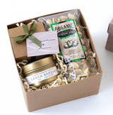 The Santa Barbara Classic Petite Gift Box features a gold tin soy candle, organic salted pistachios, and chocolate truffles in a clear bag with a green ribbon, all arranged on crinkle paper.