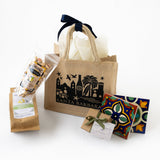 The Santa Barbara Getaway Tote gift set includes a burlap tote, snack mix, olive oil, two colorful ceramic tile coasters, and a small green-ribbon gift box, all attractively displayed on a white background.
