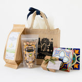 The Santa Barbara Getaway Tote gift set includes a tote bag, organic pistachios, olive oil sea salt crackers, a blue ribboned box, and two ceramic tile coasters, all elegantly arranged on a white background.