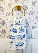 Santa Barbara Linen Tea Towel with blue toile pattern hangs on a gold ring against floral wallpaper near a light switch.