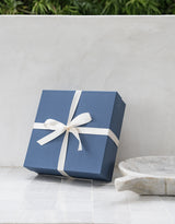 The Santa Barbara Simplicity Gift Box, blue with a white ribbon bow, sits on a white tiled surface beside a gray stone dish and vibrant leafy greens in the background.