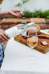 A person pours Margerum Rosé wine into a glass on a white tablecloth, with bread and condiments nearby—perfect for pairing with the Santa Barbara Wine Picnic Basket at your next beach outing.