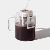 A clear glass mug holds dark coffee as a Single-Serve Pour Over Churro Coffee filter attaches to the rim, with hot water poured over Vietnamese Origin grounds. The scene is set against a plain white background with a right-side shadow.