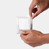 Two hands peel open a small white packet to reveal a square Single-Serve Pour Over Churro Coffee patch—perfect for enjoying Vietnamese coffee—against a plain, light background.