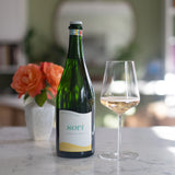A bottle of Sparkling White Non-Alcoholic Wine rests on a marble countertop next to a wine glass filled with this low sugar, dry style drink. In the background, orange roses in a vase and blurred decor set the scene.