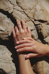 Two hands with neatly manicured nails rest on a sunlit rock, their moisturized skin glowing after applying Circular Living’s Sunset Body Oil | Clary Sage & Vetiver, evoking relaxation as sunlight highlights the contours of both hands and stone.