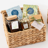 The Sweet Relaxation Gift Basket includes a woven basket with tea packets, Citrus Bath Salts, a jar of Ojai Valley Orange Honey, a brown box, and a lavender-scented soap bar arranged on shredded paper.