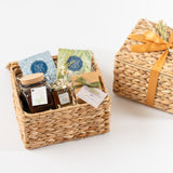 The Sweet Relaxation Gift Basket features a lidded woven basket with an orange ribbon, filled with Ojai Valley Orange Honey, soap, tea, Pink Himalayan Sea Salt, and assorted small gifts—all arranged on a white background.