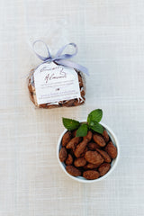 A small bowl of chocolate-covered almonds with Pink Himalayan Sea Salt, topped with mint, rests on a textured cloth. Behind it is a labeled clear bag from the Sweet Relaxation Gift Basket, tied with a lavender ribbon.