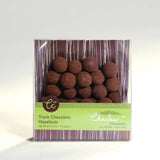 A clear box of Triple Chocolate Hazelnuts displays chocolate-coated Oregon hazelnuts inside. The green and brown packaging is marked all natural and non-GMO, making it ideal for gourmet snack lovers.