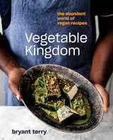 Cover of the book, Vegetable Kingdom. A person in a blue apron holds a rustic plate of tempeh over greens with seeds and sauce.