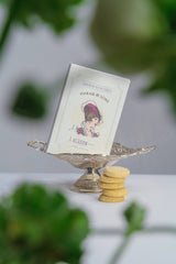 A decorative tin of Verdon Lavender Shortbread Cookies, made by Makabi & Sons in California, sits in a silver dish beside a stack of round cookies, with greenery softly blurred in the foreground.