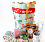 A gift set featuring the Views of San Francisco Tote Bag (natural cotton canvas), ribbon, snacks, a white tumbler, vintage illustration postcards, pink treat box, green tin, and breadsticks—all arranged neatly on a white surface.