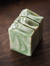 Three rectangular bars of White Sage Ojai Botanika Soap, marbled green and cream, stand upright on a dark wooden surface. Each vegan soap bar is topped with a sprinkling of dried herbs.
