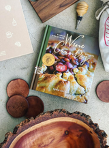 The Wine Country Cookbook rests on a stone surface, surrounded by wooden coasters, a bowl, a wine bottle stopper, and part of a cloth bag—an ideal companion for wine country cooking and tasting.