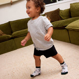 Smiling toddler in a gray T-shirt and black shorts walking across a living room rug in Kids Socks that Protect Bears
