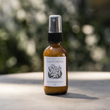 Amber spray bottle labeled California Room & Body Mist on a table outdoors, clean scent, blurred greenery in background.