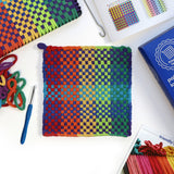 Finished rainbow woven potholder beside colorful cotton loops, a crochet hook, and the PRO loom kit box
