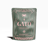 A sage green stand-up pouch of GATO Dark Chocolate Covered Dates in pistachio butter flavor, with a pink palm tree illustration and a pink and green checkered border along the bottom.