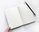 A Linen Hardcover Journal lies open on a white surface, with a black pencil resting on its right page.