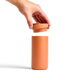 A hand lifts the lid off a Ceramic Coated Travel Mug in Terracotta against a white background.