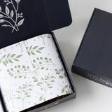 Golden State Gift Basket with leafy paper, dark lid, and artisan snacks, partly open on a white surface.