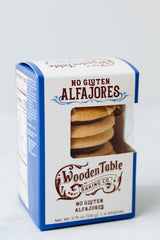 A blue and white box of Alfajores Gluten-Free Cookies with a clear window displays stacked treats. The packaging has brown decorative text and designs, highlighting dulce de leche-filled gluten-free shortbread cookies.