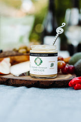 A jar of Artichoke and Parmesan Tapenade sits on a wooden board with cheese, grapes, red peppers, and a decorative spoon—creating an inviting appetizer spread, with wine glasses softly blurred in the background.