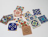 A set of Assorted Ceramic Tile Coasters (Bulk) featuring vibrant handcrafted patterns is displayed on a white surface. One coaster reveals a cork backing stamped with Santa Barbara Company, reflecting Spanish heritage decor.