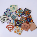 A variety of colorful, patterned Assorted Ceramic Tile Coasters (Bulk) are arranged on a white surface, displaying geometric, floral, and abstract Spanish-inspired designs. One tan coaster in the corner is labeled The Santa Barbara Company.