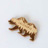 The California Bear Wood Magnet, made from sustainably harvested wood and laser-engraved with mountains, pine trees, and "California," is displayed on a white background.