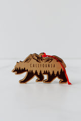 The California Bear Wood Ornament is crafted from sustainably harvested wood, engraved with mountains, trees, and “California,” and comes with a red ribbon loop for hanging.