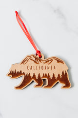 The California Bear Wood Ornament is crafted from sustainably harvested wood, featuring engraved mountain and tree designs, hangs from a red ribbon, and is displayed on a white marble surface.