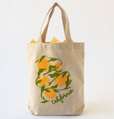 The California Oranges Organic Cotton Tote is a beige canvas bag with handles, decorated with California oranges, green leaves, white blossoms, and “california” in script at the bottom. Yellow paper peeks from the top.