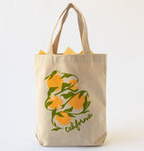 The California Oranges Organic Cotton Tote is a beige canvas bag with handles, decorated with California oranges, green leaves, white flowers, and "California" on the front. Yellow tissue paper peeks out from the top.