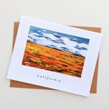 The California Poppy Field Note Card showcases a vibrant California landscape with orange poppy fields, green accents, and a blue sky. "California" is printed below the image. Comes with a brown envelope—ideal for gifting or personal notes.