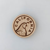 The California Quail Wood Magnet is a round, sustainably harvested wood magnet with a light background, featuring an engraved California quail and the word "CALIFORNIA.