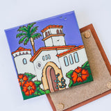 The Courthouse of Santa Barbara Art Trivet is a decorative ceramic tile featuring a hand-painted, colorful scene of the Santa Barbara County Courthouse with red-tiled roofs, arches, palms, red flowers, and cork pads on the brown base.